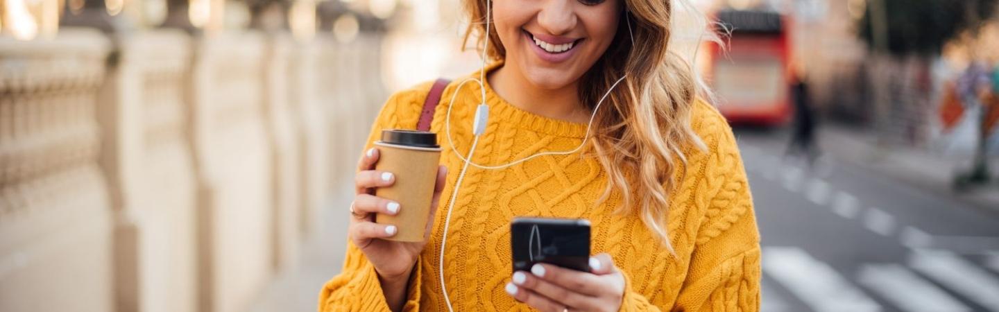 Image shows a woman walking along a London street with a cup of coffee, headphones in and a mobile phone in her hand. She is smiling at her phone. 
